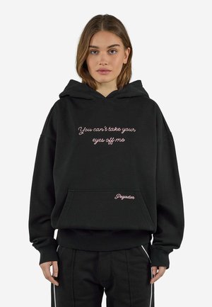 Kvinde iført sort hættetrøje med pink tekst "You can't take your eyes off me" og "Pegador" på frontlommen, stående mod en hvid baggrund.