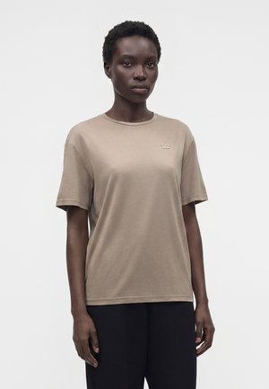 Sort kvinde med kort hår iført en beige t-shirt med korte ærmer og sorte bukser, stående mod en ensartet lys baggrund.