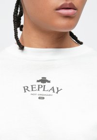 Κοντινό ενός ατόμου που φοράει λευκό πουκάμισο με μαύρο κείμενο που γράφει "REPLAY NOT ORDINARY 1981" κάτω από το λαιμό.