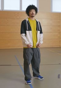 Outfit sportivo con una giacca nera e bianca sopra una maglietta gialla brillante, abbinata a pantaloni cargo grigi larghi e sneaker blu.