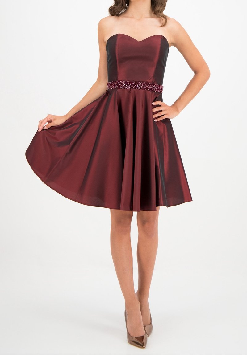 Robe bustier en satin bordeaux avec une ceinture perlée, jupe évasée longueur genoux, portée avec des escarpins métalliques à bout pointu.