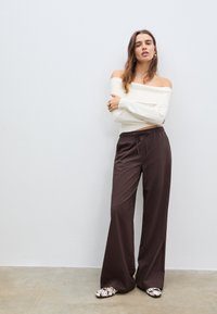 Off-Shoulder-Rippstrickoberteil in Weiß, braune weit geschnittene Hose mit Kordelzug und gemusterte Slipper, vor einem neutralen Hintergrund.