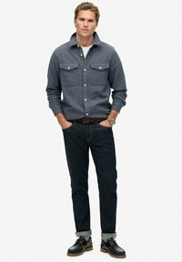 Niebieska, pleciona koszula z dwiema kieszeniami na piersi i guzikami, w połączeniu z ciemnoniebieskimi dżinsami i czarnymi sznurowanymi butami, z podwiniętymi mankietami.