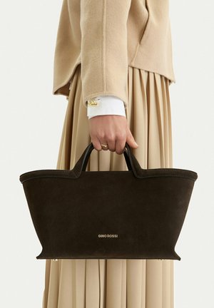 Hand hält eine schwarze Gino Rossi Wildleder-Shopper-Tasche, trägt einen beigen Mantel und einen plissierten beigen Rock, goldener Ring und Manschettenknopf sichtbar.