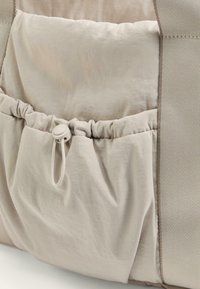 Bež tkaninasta torbica z nabranim zgornjim delom, zapiranjem na vrvico in gladko teksturo. Robustni šivi in kontrasten teksturiran odsek na strani.
