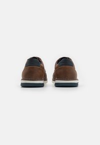 Chaussures en cuir marron avec un panneau textile bleu marine au niveau du talon, semelle blanche et bleu foncé, et couture blanche le long de la semelle intermédiaire.