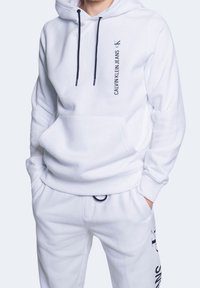 Sudadera con capucha blanca y pantalones de chándal hechos de tela suave. La sudadera tiene cordones negros y un pequeño logo en el pecho y en la pierna del pantalón.