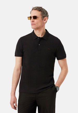 Homme portant un polo noir à manches courtes, un pantalon noir, des lunettes de soleil teintées de jaune et une montre-bracelet, regardant sur le côté avec une main dans la poche.