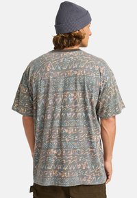 Camisa de manga corta en tejido verde apagado con patrones abstractos multicolores, que incluyen olas y espirales, con un ajuste relajado y cuello redondo.