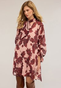 Blumenkleid aus zartem rosa Stoff, das mit großen roten Blumenmustern, einem hohen Kragen mit einer Schleife und weit schwingenden Ärmeln ausgestattet ist.