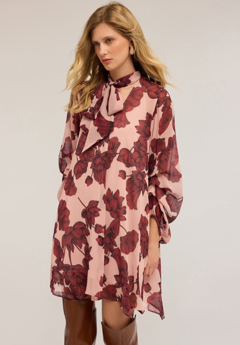 Blumenkleid aus zartem rosa Stoff, das mit großen roten Blumenmustern, einem hohen Kragen mit einer Schleife und weit schwingenden Ärmeln ausgestattet ist.