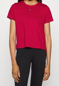 Short-sleeve, cropped t-shirt in vibrant red, made of textured fabric, featuring a small logo accent in the center. Paired with black leggings.