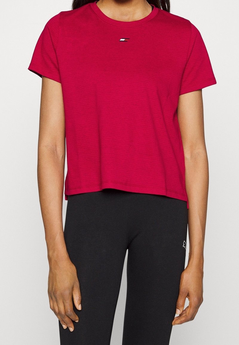 Short-sleeve, cropped t-shirt in vibrant red, made of textured fabric, featuring a small logo accent in the center. Paired with black leggings.