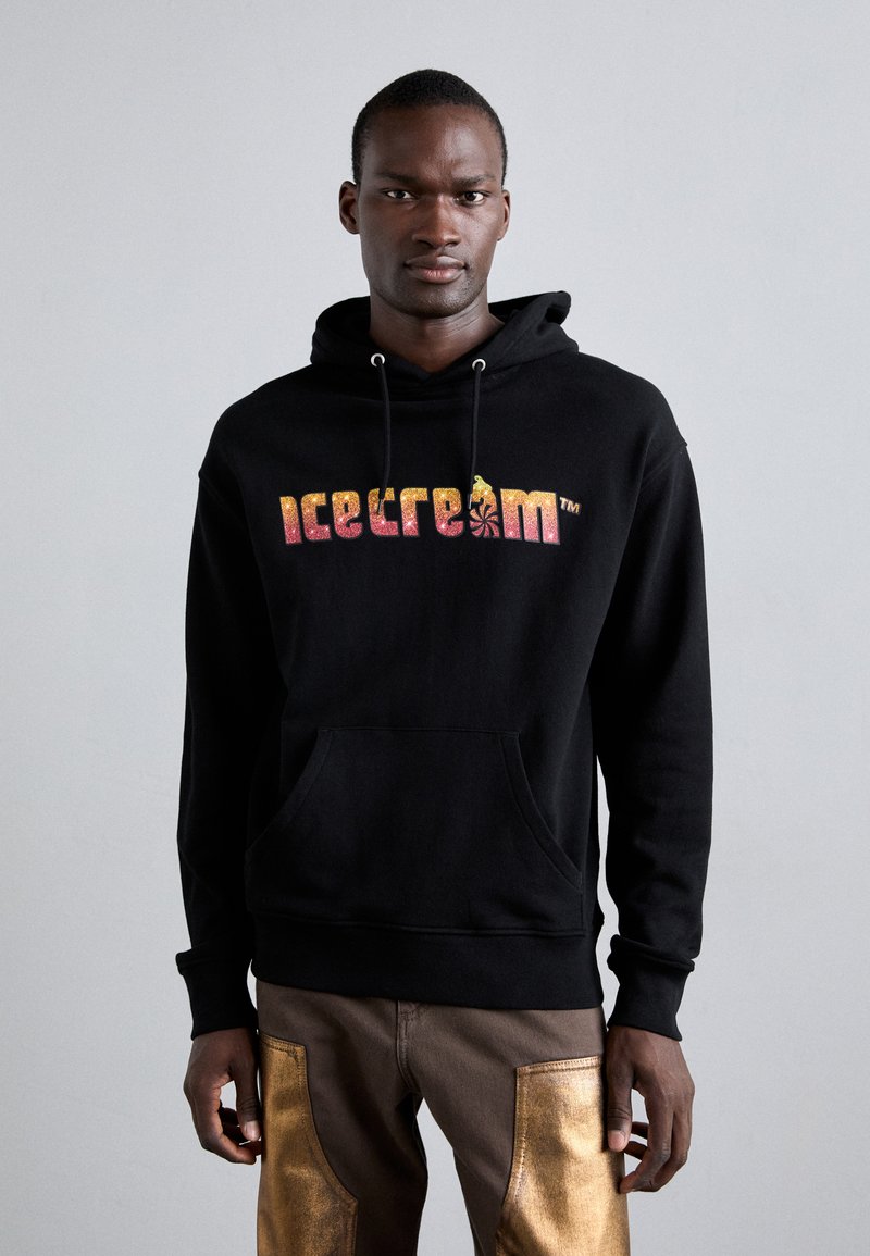 Haut à capuche noir en tissu à texture douce. Présente un logo coloré "glace à la crème" et une poche avant. Associé à un pantalon marron avec des accents dorés.