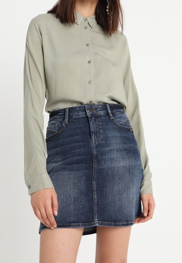 Femme portant une chemise à manches longues boutonnée vert clair rentrée dans une mini-jupe en denim bleu foncé taille haute.