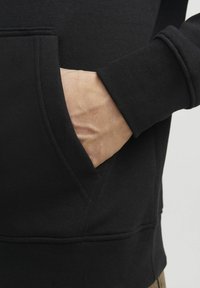 Sort hoodie med en kengurulomme, ribbede manchetter og en blød tekstur. Nærbillede der fremhæver syninger og lomme detaljer.