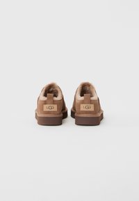 Pari mokasin suède UGG cokelat muda dengan lapisan bulu lembut, dilihat dari belakang, di latar belakang abu-abu terang yang polos.