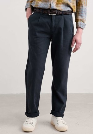 Donkerblauwe, geplooide broek met een relaxte pasvorm, gemaakt van lichtgewicht stof, voorzien van een gevlochten leren riem in bruin, gecombineerd met crèmekleurige schoenen.