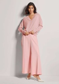 Helle rosa gestrickte Sweatshirt mit V-Ausschnitt und langen Ärmeln, kombiniert mit weiten rosa Hosen. Das Schuhwerk hat spitze Zehen und Absatzakzente.