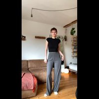 Camiseta negra corta con mangas cortas, combinada con pantalones de pierna ancha gris de cintura alta. El entorno cuenta con un sofá marrón y plantas de interior.