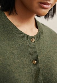 Zöld kötött kardigán, kerek nyakkivágással, bordázott mandzsettákkal és két barna gombbal az elülső záródásért. Puha textúrájú és laza stílusú.