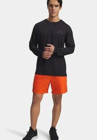 Maglietta nera a maniche lunghe, pantaloni sportivi arancioni e scarpe nere. La maglietta ha un logo discreto; il tessuto appare leggero e traspirante.