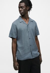 Άνδρας με σκούρο δέρμα και κοτσίδες που φοράει μπλε πουκάμισο με κοντά μανίκια και μαύρο παντελόνι, κοιτάζοντας προς τα πλάγια με φόντο λευκό.