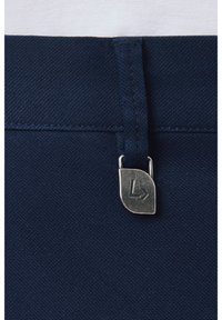 Granatowy materiał pasa o gładkiej fakturze, z metalową metką logo przymocowaną metalowym zapięciem. Zbliżenie uwydatnia szwy.