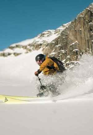 Een skiër in een geel-zwarte jas daalt een besneeuwde helling af, draagt een zwarte helm en skibril, terwijl hij een sneeuwspray creëert tegen een rotsachtige achtergrond.