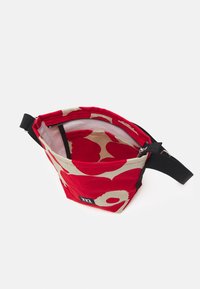 Rød stoftaske med et beige blomster mønster, toppen med lynlås lukning, sort rem og et lille logo tag på siden. Indersiden er foret.