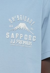 Ανοιχτό μπλε βαμβακερό μπλουζάκι με ανάγλυφο λευκό σχέδιο αντίθεσης που απεικονίζει βουνά και κείμενο που γράφει "90th Brigade Sapporo JJ Premium".