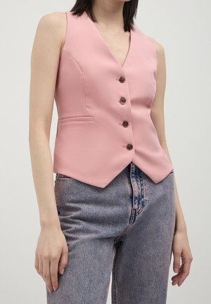 Femme portant un gilet rose sans manches boutonné associé à un jean bleu délavé taille haute sur un fond uni.