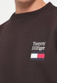Brun genser med ribbet rund hals. Har et brodert logo med teksten "Tommy Hilfiger" og et rektangulært tri-farge emblem.