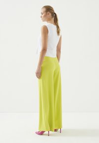 Witte, geribbelde, mouwloze top gecombineerd met hooggewaist, wijde, felgroene broek en roze hoge hakken. Model gezien vanaf de achterkant.