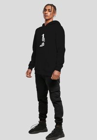 Jeune personne avec des nattes portant un sweat à capuche noir avec un motif d'oiseau et un imprimé de baskets, ainsi que un pantalon cargo noir et des baskets montantes noires.