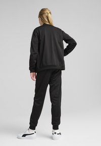 Veste noire avec des accents latéraux blancs, associée à un pantalon de jogging noir. Le matériau semble lisse et athlétique, complété par des baskets blanches.