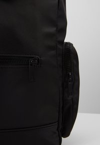Mochila negra con tela suave, cremalleras prominentes y un bolsillo lateral con cierre de cremallera. Diseño simple con una superficie texturizada.