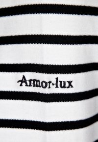 Stribet bomuldsstof i sort og hvid med "Armor-lux" broderet i sort. Nærbillede fremhæver tekstur og design.