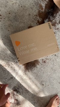 Rjava kartonska škatla z oranžnim logotipom, ki vsebuje natisnjen besedilo: "Ljubite me. Nosite me." Škatla kaže rahlo poškodbo na vogalu, postavljena na teksturirano površino.