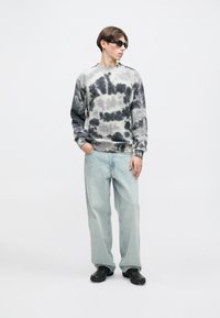 Bluza tie-dye w szaro-czarnych odcieniach, zestawiona z jasnoniebieskimi szerokimi dżinsami i czarnymi trampkami, dopełniona okularami przeciwsłonecznymi.