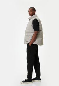 Lys grå polstret vest med høj krave, lynlåslukning og forlommer, båret over en sort skjorte, parret med sorte bukser og sneakers.