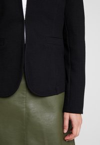 Personne portant un blazer noir texturé sur un haut blanc et une jupe en cuir vert, avec sa main droite relaxée à ses côtés.