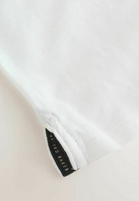 Camiseta de algodón blanca con un escote redondeado, costuras visibles y una etiqueta negra con "by TED BAKER" en el dobladillo.