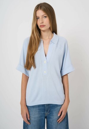 Jeune femme aux longs cheveux blonds portant une chemise à manches courtes bleu clair avec col et un jean bleu, debout devant un fond uni.