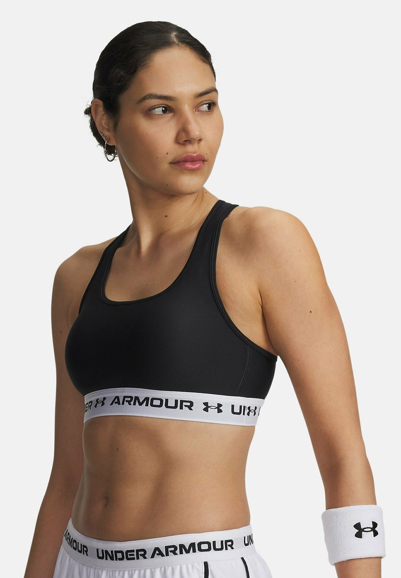 Sujetador deportivo negro con un diseño ajustado y una banda ancha con el logo blanco de Under Armour. La tela parece suave con una textura elegante.