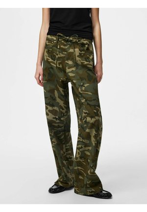 Persona che indossa pantaloni mimetici larghi con coulisse in vita, canotta nera e scarpe basse nere, in piedi davanti a uno sfondo semplice.