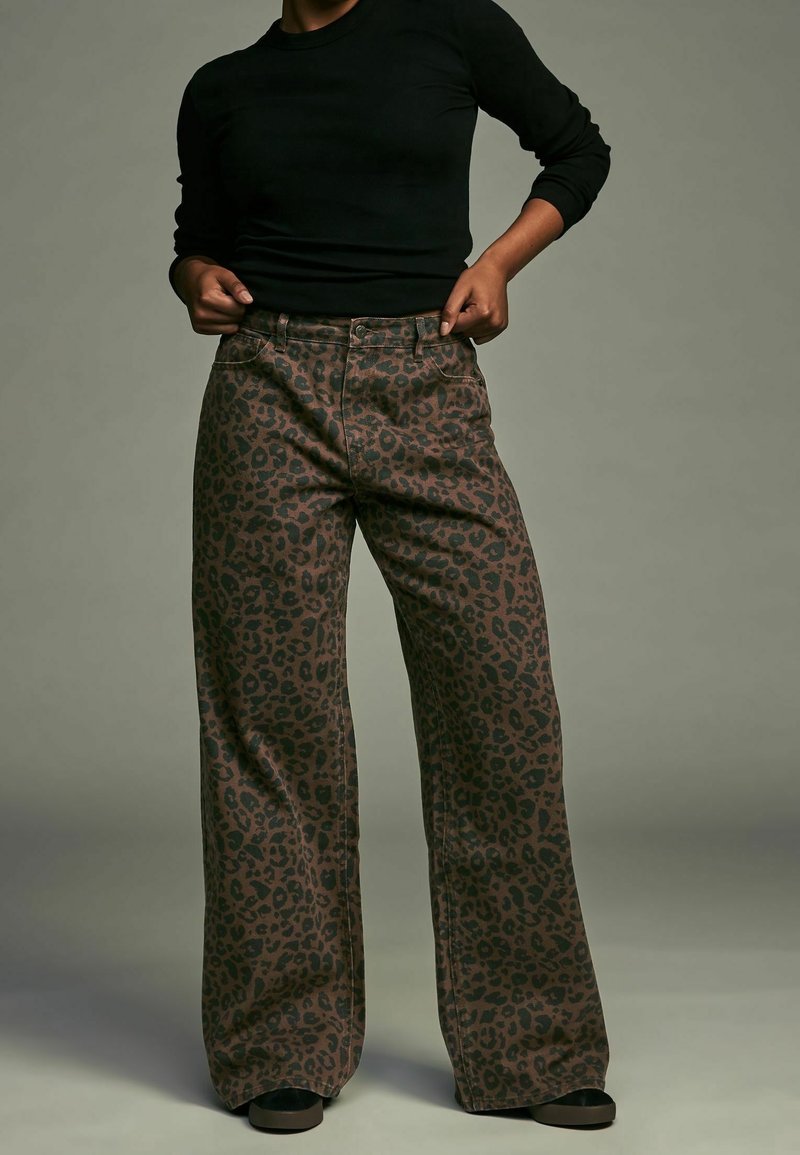 Personne portant un pantalon marron à jambes larges avec un imprimé léopard noir et une chemise noire à manches longues sur un fond uni.