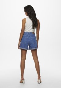 Pantalones cortos de mezclilla en azul claro con cintura alta, dobladillos enrollados y dos bolsillos traseros. Combinados con una camiseta sin mangas blanca y sandalias de tacón cuadrado beige.