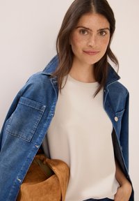 Ung kvinde med brunt hår og fregner bærer en blå denimjakke over en lys beige skjorte og holder en tan ruskinds taske.
