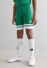 Barn som bär gröna Adidas sportshorts, vita knähöga strumpor med svart logotyp och vita fotbollsskor som står på en grå golv.